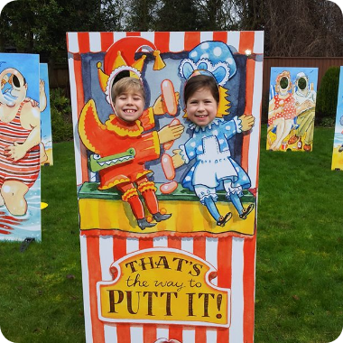 Two young children posing behind a red-and-yellow Punch and Judy photo cutout board outdoors.