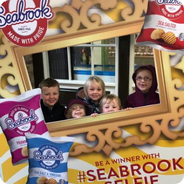 Promotional graphic featuring children posing behind an ornate gold frame cutout, surrounded by Seabrook Crisps branding and packet images.